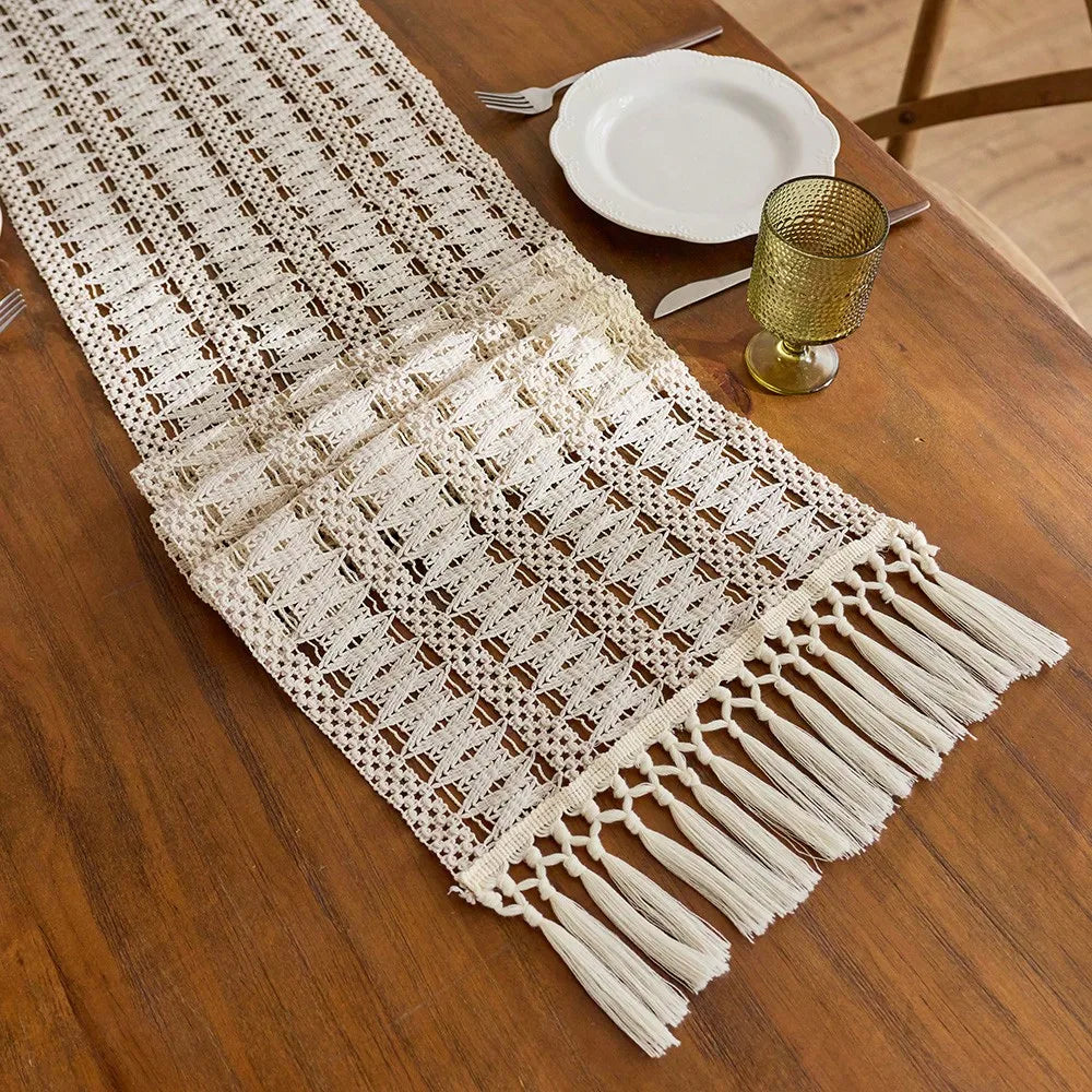 Beige bohemian farmhouse table runner with fringe detail placed on a wooden table next to a gold goblet and silverware.