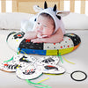 A baby wearing a cow-themed hat is lying on a high contrast tummy time pillow with various animal-themed teething rings.
