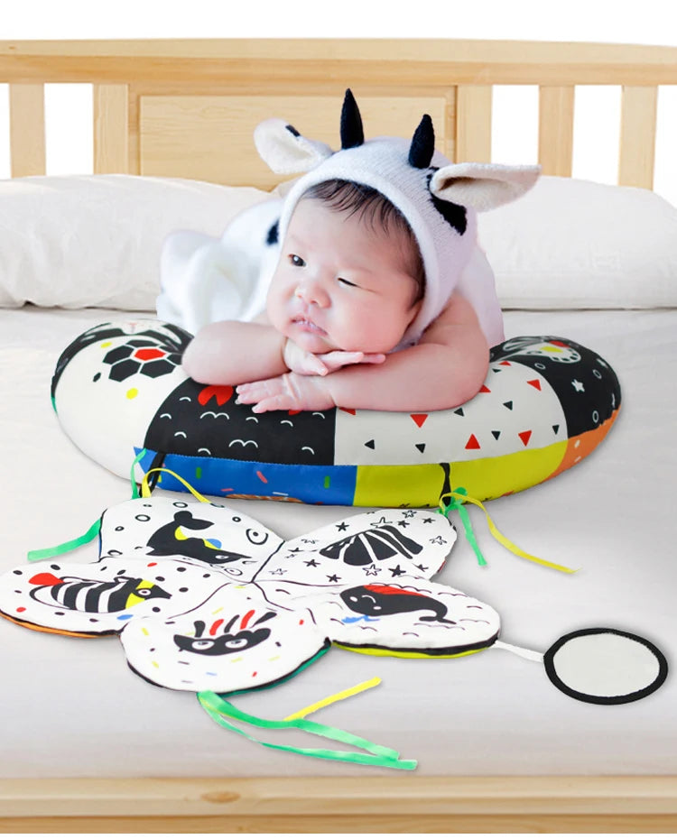 A baby wearing a cow-themed hat is lying on a high contrast tummy time pillow with various animal-themed teething rings.