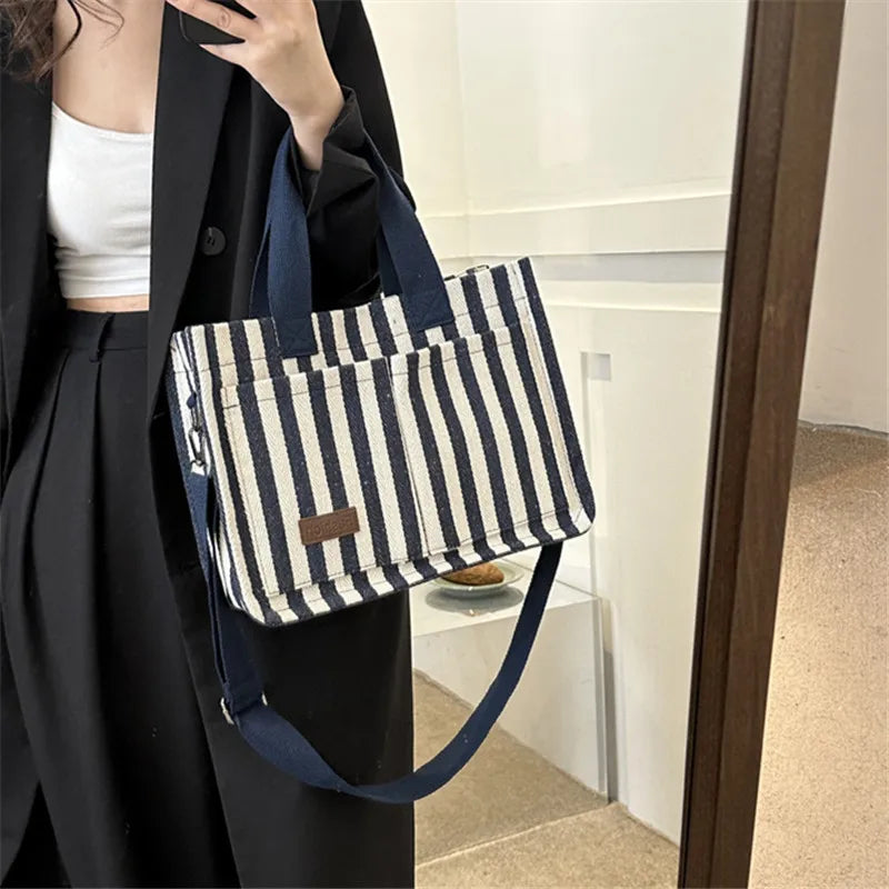 A woman holds a striped canvas tote bag with navy straps and inner pockets against a black coat.
