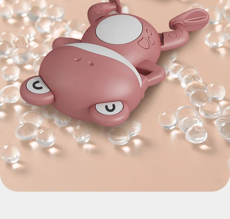 A pink wind-up swimming frog bath toy floating among clear bubbles on a light surface.