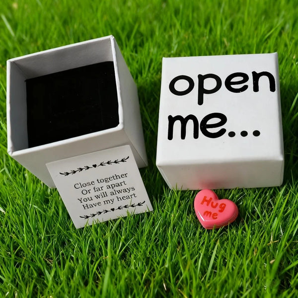 A white gift box with the text open me... and a pink heart charm resting on green grass.