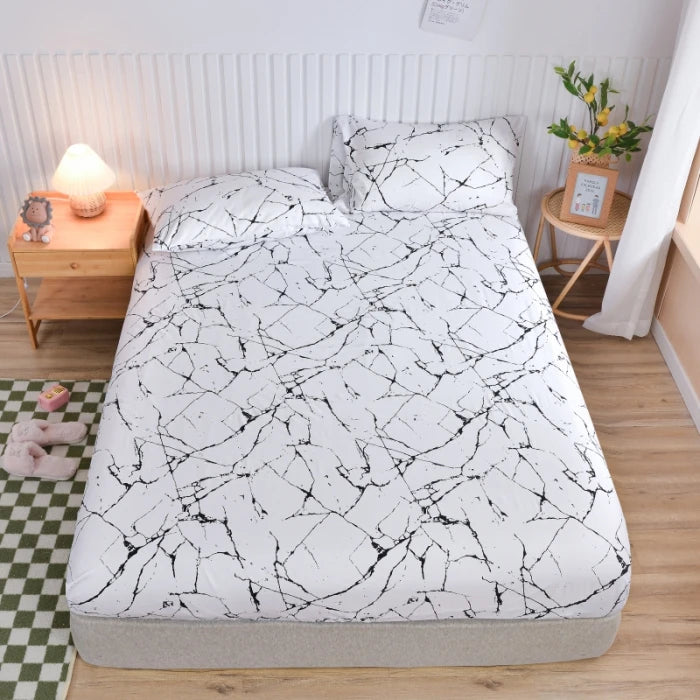 A white fitted bed sheet with a black marble print is displayed on a bed, next to a bedside lamp and wooden furniture.