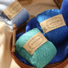 A collection of blue and teal pure cotton yarn balls are displayed in a woven basket.