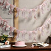 Pink sheer ribbon bowknot banner garland displayed against a light wall with a cake and book.