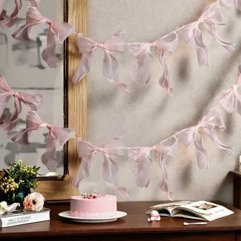 Pink sheer ribbon bowknot banner garland displayed against a light wall with a cake and book.