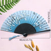 A light blue folding hand fan decorated with white floral patterns and a black handle, shown against a pink background.