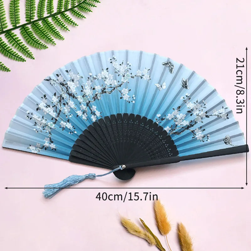 A light blue folding hand fan decorated with white floral patterns and a black handle, shown against a pink background.