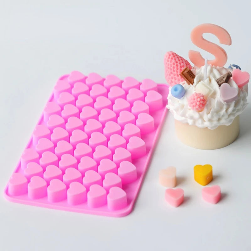 A pink silicone mold tray filled with heart-shaped silicone molds, next to a decorative cake topper featuring an 'S' and vari