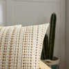 A close-up of a cream and tan woven linen pillow cover with a textured pattern, set against a blurred background of green cac