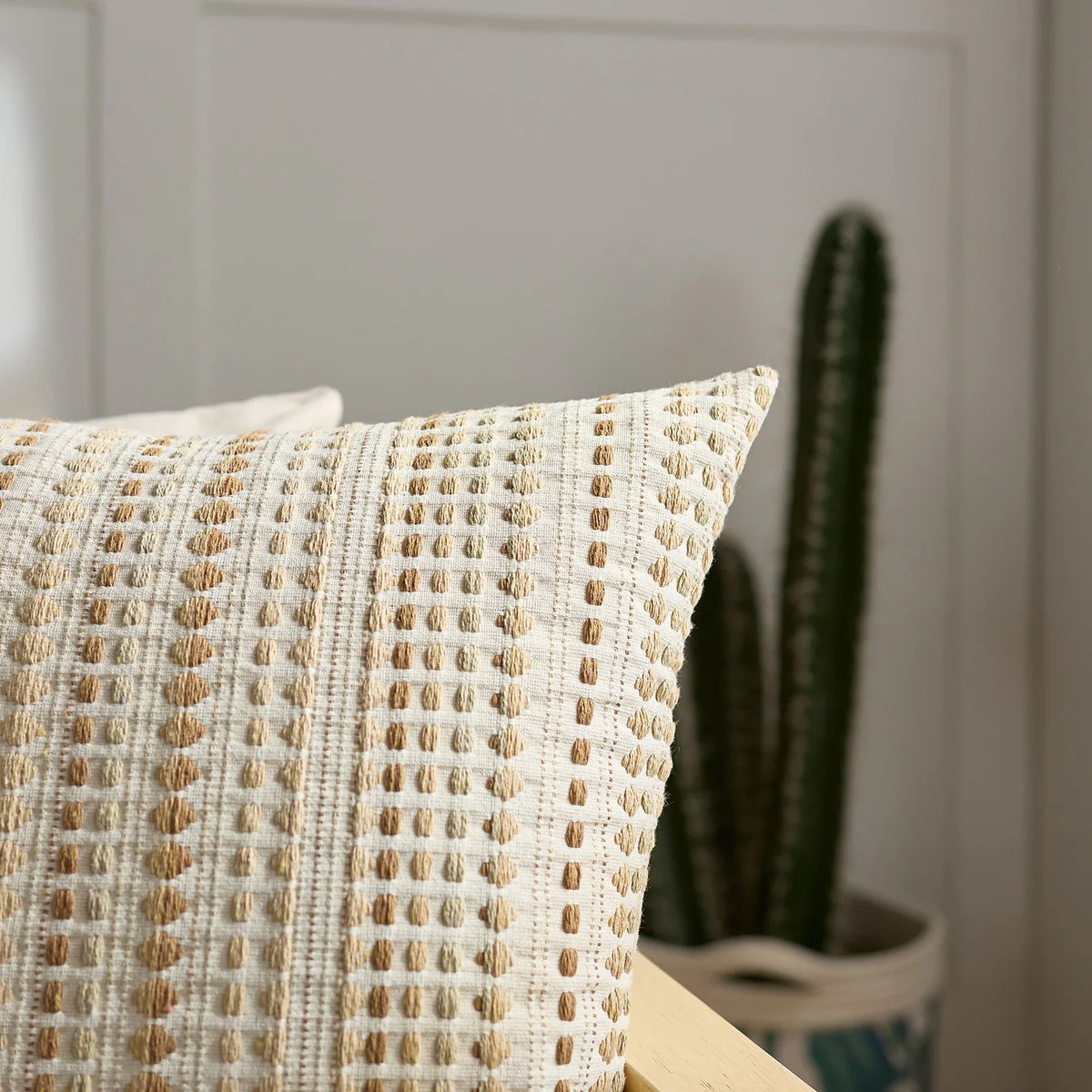 A close-up of a cream and tan woven linen pillow cover with a textured pattern, set against a blurred background of green cac