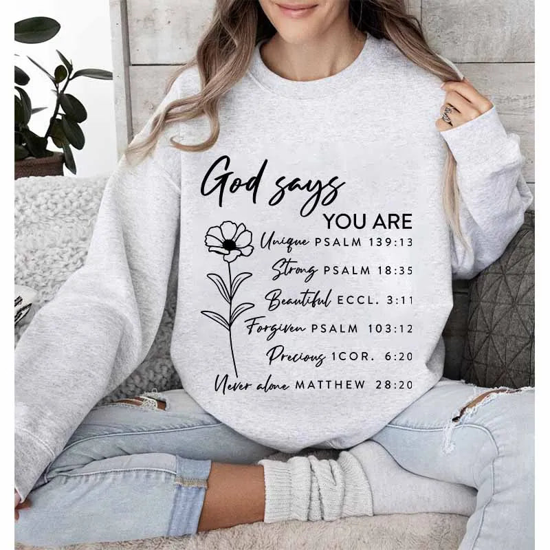 A woman wearing a light gray sweatshirt with black text that reads God says you are unique strong beautiful forgiven precious