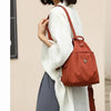 A woman wearing a light beige jacket and dark pants carrying a burnt orange mini backpack.