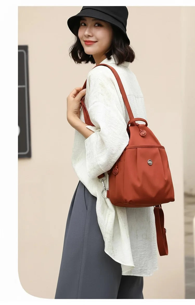 A woman wearing a light beige jacket and dark pants carrying a burnt orange mini backpack.