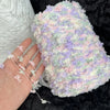 A hand is holding a fluffy, variegated yarn blend in pastel pink, purple, and green tones.