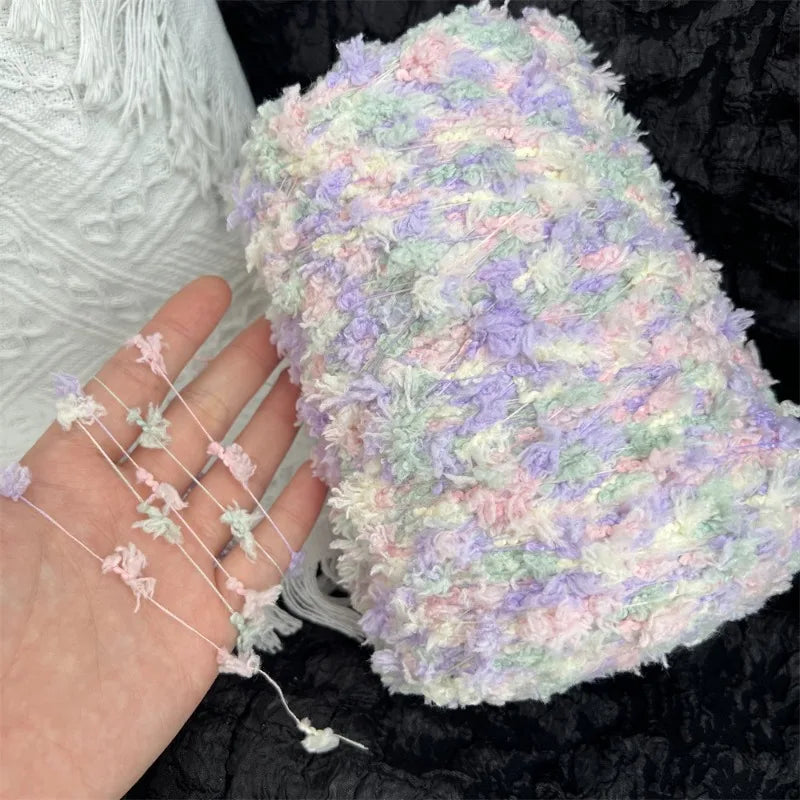A hand is holding a fluffy, variegated yarn blend in pastel pink, purple, and green tones.