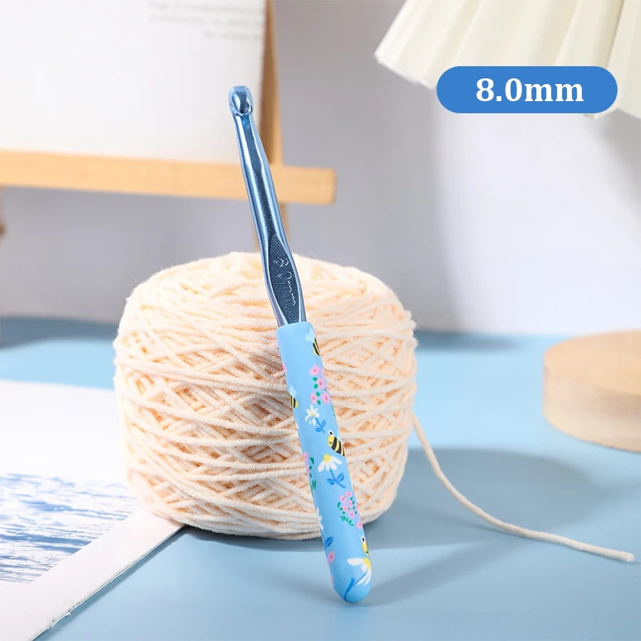 A blue ergonomic crochet hook with a bee design is shown next to a ball of light pink yarn.
