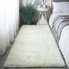 A fluffy white silk-touch area rug placed on a wooden floor next to a bed with gray patterned bedding.