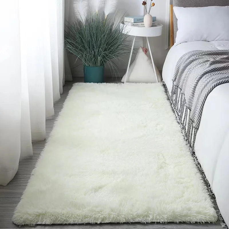 A fluffy white silk-touch area rug placed on a wooden floor next to a bed with gray patterned bedding.