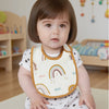 A baby wearing a white bib with a rainbow design and a brown trim sitting on a light surface.