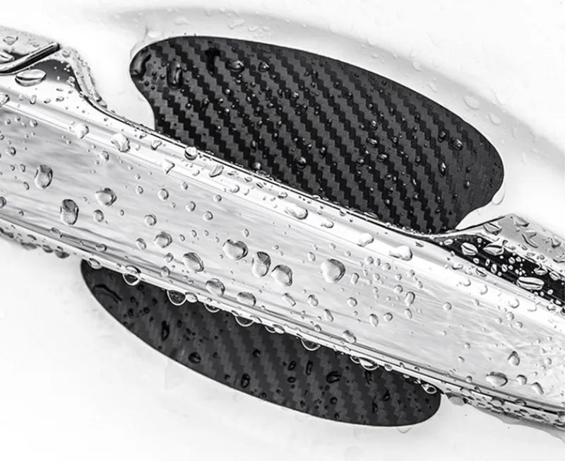 Close-up of a carbon fiber car door handle protected by a clear film, showing water droplets on the surface.