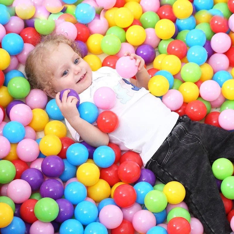 A toddler is smiling while surrounded by a large pile of colorful plastic ball pit balls.