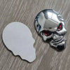 A chrome skull emblem badge featuring a red eye, placed next to its white backing.