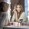 A woman applies makeup in front of a mirror with a waterproof neoprene cosmetic bag on the counter.