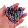 A hand holds a black embroidery patch with purple and green stitching that reads Love Conquers All Even Thorns
