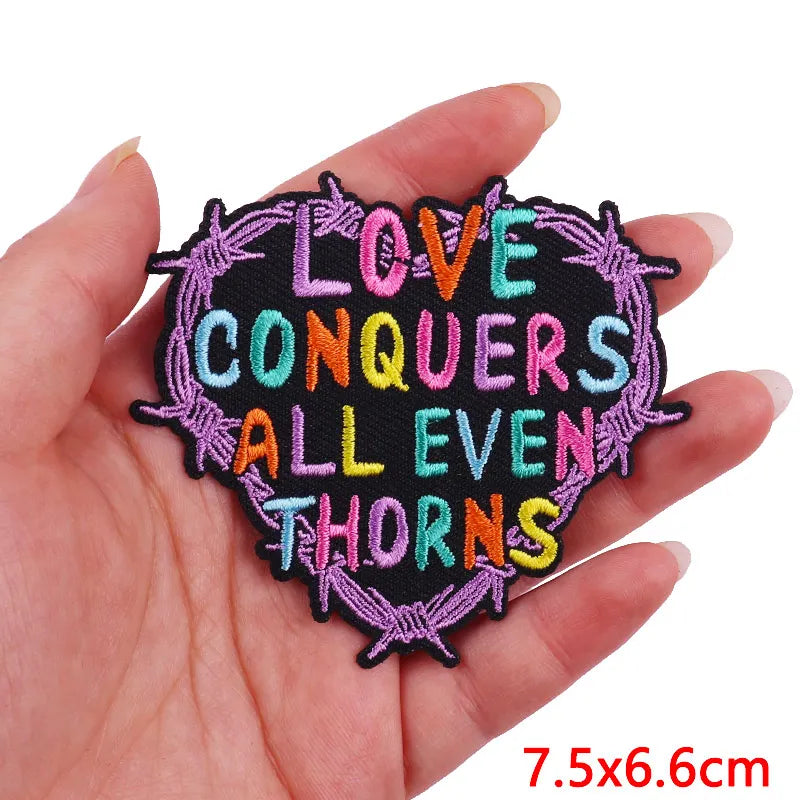 A hand holds a black embroidery patch with purple and green stitching that reads Love Conquers All Even Thorns