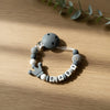 A gray silicone pacifier clip bracelet featuring a custom name engraved on the beads, resting on a wooden surface.