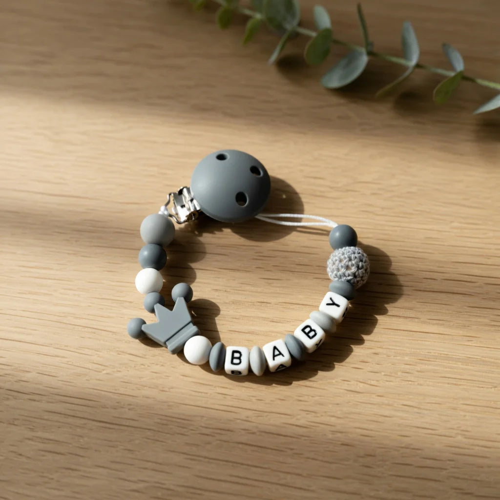 A gray silicone pacifier clip bracelet featuring a custom name engraved on the beads, resting on a wooden surface.