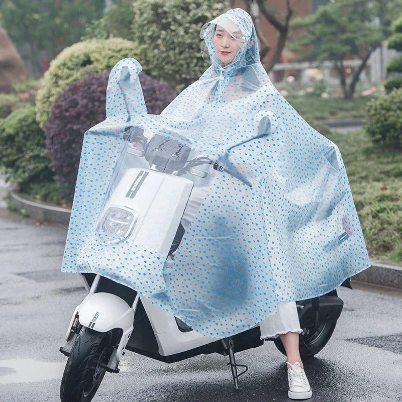 A woman wearing a transparent blue polka-dot waterproof motorcycle rain poncho with a hood, standing next to a white scooter.