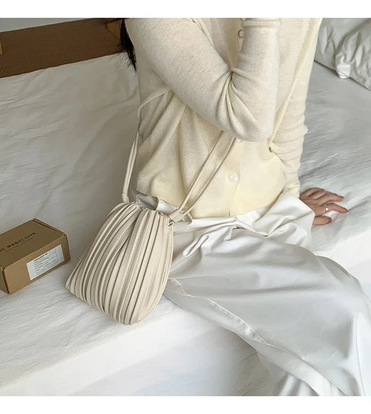 A person wearing a cream-colored top and white pants holds an elegant pleated faux leather crossbody bag.