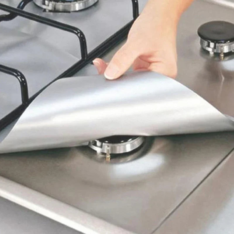 A hand places a silver reusable square gas hob protector liner onto a stovetop burner.
