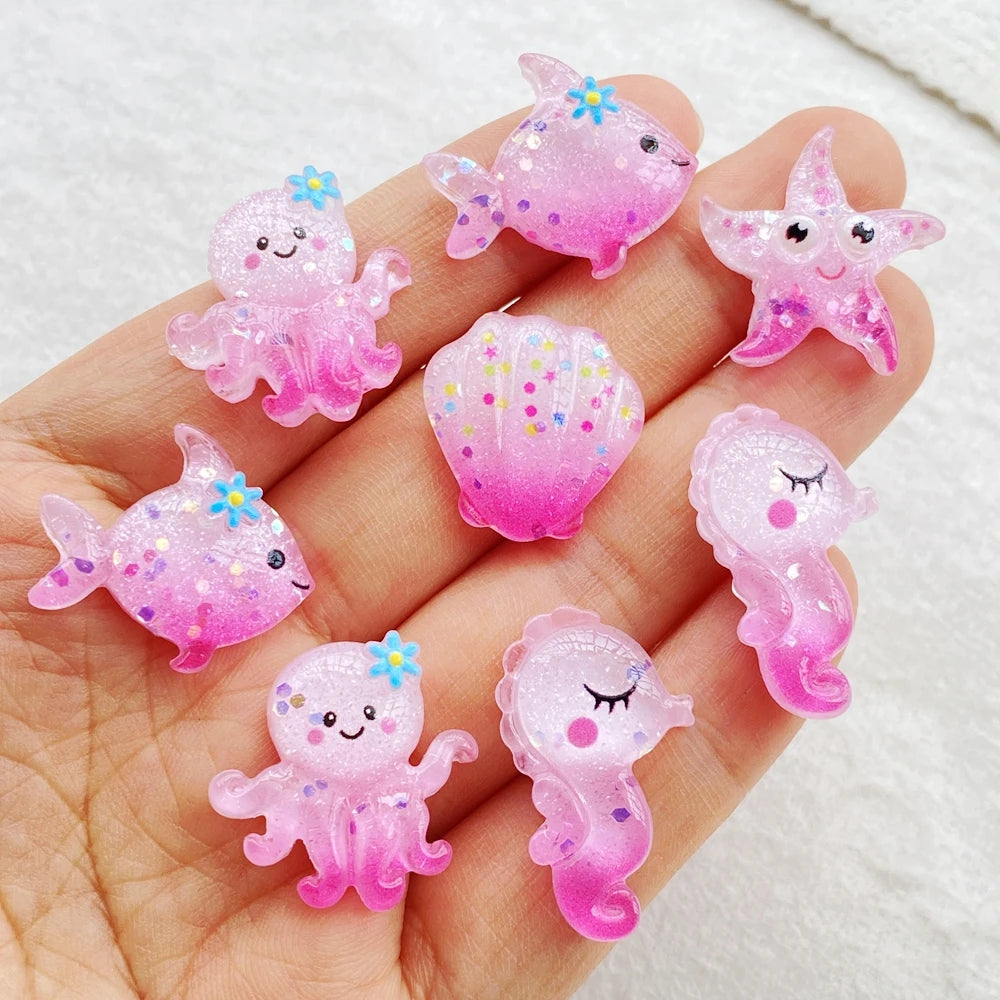 A hand holds several pink and purple glittery resin charms shaped like sea creatures for crafting.