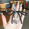 A hand holds a black beaded pearl bow keychain with a silver heart charm.