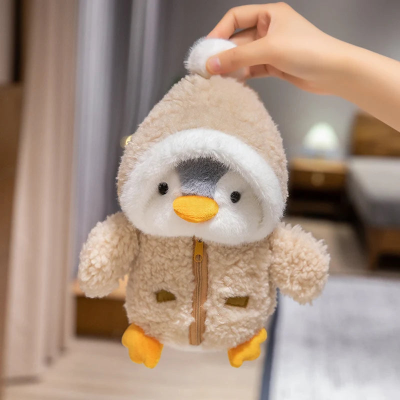 A hand is holding a fluffy, light brown penguin plush toy with a white hat and yellow feet.