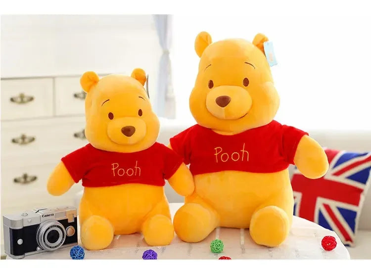 A yellow Winnie the Pooh plush toy bear wearing a red shirt sitting next to a camera.