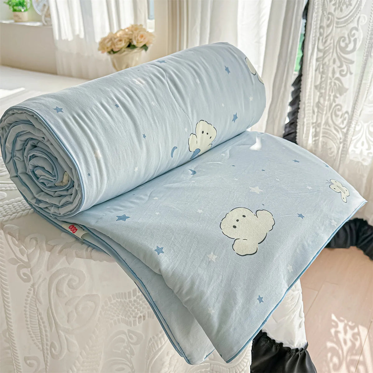 Light blue quilt with white cloud and star patterns rolled up on a white surface.