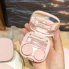 A pink compact contact lens travel case is shown open, displaying clear lenses and round storage compartments.