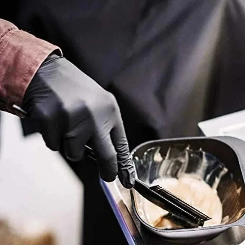 A hand wearing a black nitrile disposable glove is using a brush to apply a substance inside a black bowl.