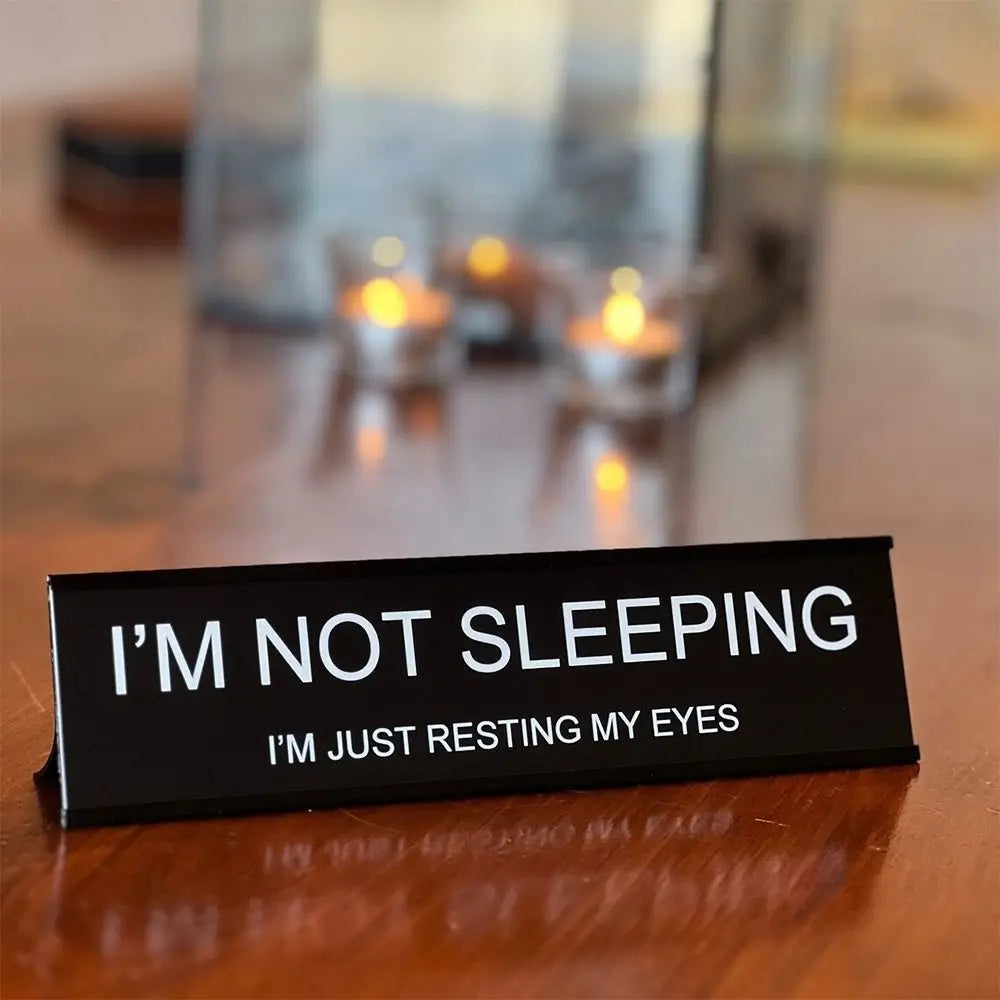 A black desk sign reads I'M NOT SLEEPING I'M JUST RESTING MY EYES placed on a wooden surface with blurred candles in the back