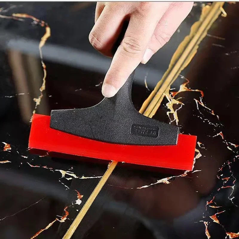 A hand is using a black silicone scraper tool to remove grout or sealant from a dark surface.