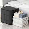 A black foldable wardrobe storage bag with a double zipper is placed next to a white bed with folded white linens.