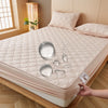 A beige waterproof mattress protector is shown on a bed, with pillows and a glass, highlighting its quilted texture.