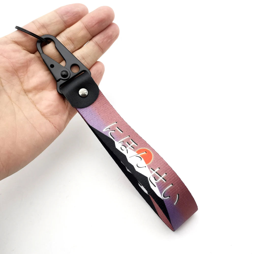 A hand holds a black and purple keychain featuring Japanese text on a strap with a steel clip.