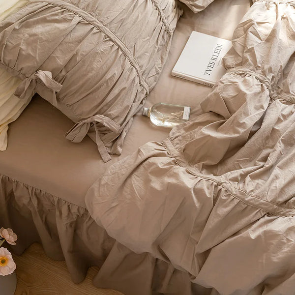 A beige washed cotton ruffle bedding set with bow ties and a glass bottle on a light surface.