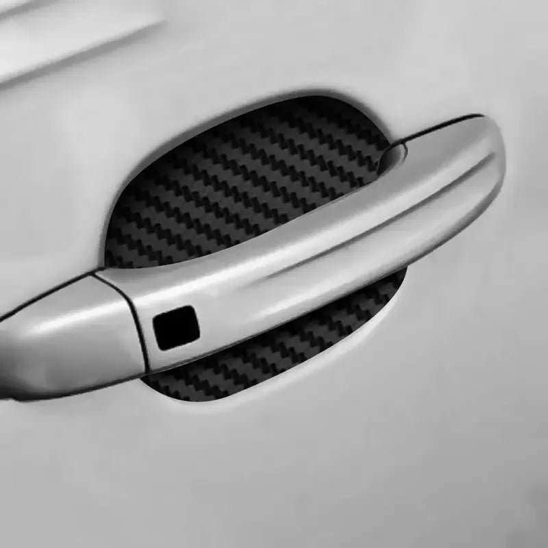 A close-up view of a silver car door handle with a black carbon fiber protective cover installed on the door.