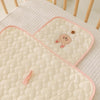 A beige quilted portable diaper changing pad with a cute brown bear design resting on a white textured surface.